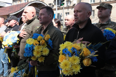 На фото зображено урочисту та водночас емоційно стриману подію на вулиці міста.

У центрі — група чоловіків, серед яких є військові та цивільні, що стоять у шерензі. Більшість із них тримають у руках букети квітів у синьо-жовтих кольорах — це поєднання нагадує кольори українського прапора. У композиціях видно жовті троянди, хризантеми та сині квіти, перев’язані стрічками.

Чоловіки виглядають серйозними та зосередженими. Деякі з них тримають праву руку на грудях — це жест поваги, який зазвичай супроводжує виконання гімну або хвилину мовчання. На задньому плані видно ще людей, зокрема військових у формі.

Подія відбувається на міській вулиці: позаду — будівлі, світлофор, а також вхід до якоїсь споруди або підземного переходу.

Загальний настрій фото — стримано-патріотичний і шанобливий, ймовірно, це церемонія вшанування, покладання квітів або пам’ятний захід.