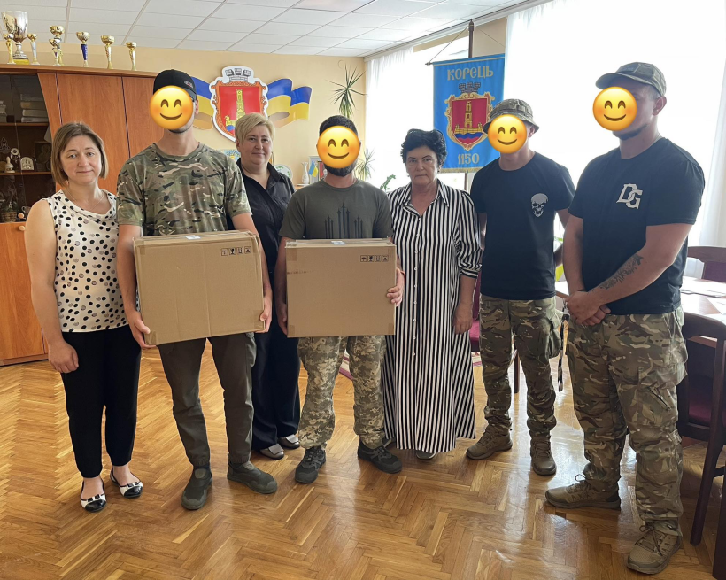 На цьому фото зафіксовано момент передачі допомоги у приміщенні з прапорами та гербом міста Корець. У центрі кадру — військовослужбовці у камуфляжній формі з закритими для публікації обличчями. Вони тримають у руках дві великі картонні коробки з технікою або спорядженням. Поруч — представники Корецької громади, які передають ці необхідні речі. Атмосфера знімка тепла та урочиста, усі учасники процесу демонструють єдність і підтримку наших захисників.