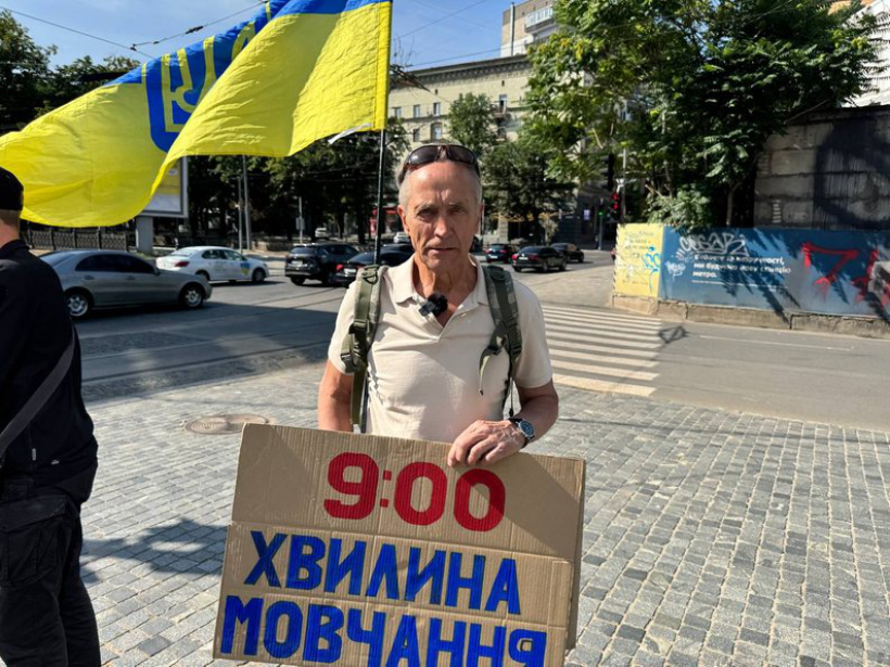 На передньому плані чоловік середнього віку, з сивим волоссям, у світлій футболці-поло й рюкзаку.
Він тримає картонну табличку з написом:
9:00 ХВИЛИНА МОВЧАННЯ (текст червоним і синім кольорами).
За його спиною розвивається великий жовто-блакитний прапор України з тризубом.
Довкола – міська вулиця: припарковані автівки, пішохідний перехід, будівлі та дерева. Атмосфера спокійна, сонячна погода.