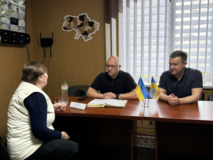На фото зображені 2 чоловіки,що сидять за круглим столом, та жінка яка ділиться своїми проблемними питаннями. На фоні вікно, а на стіні висить карта України