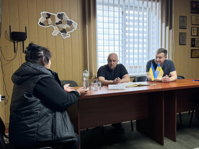 На фото зображені 2 чоловіки,що сидять за круглим столом, та жінка яка ділиться своїми проблемними питаннями. На фоні вікно, а на стіні висить карта України