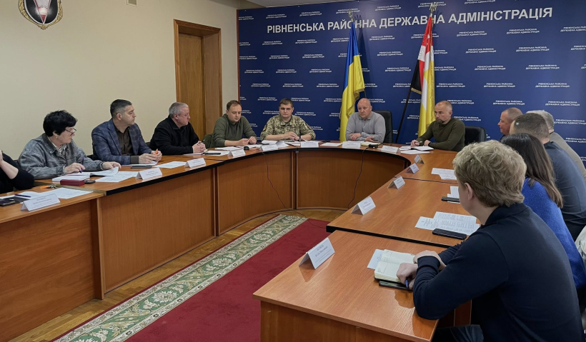 На фото зображено групу людей, які сидять за столом переговорів. На фоні синя вивіска з назвою огранізації та прапори України і м. Рівне
