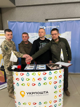 На фото зображено 4 чоловіки на синьому фоні, вони стоять біля столу з вивіскою назви і логотипу Укрпошти