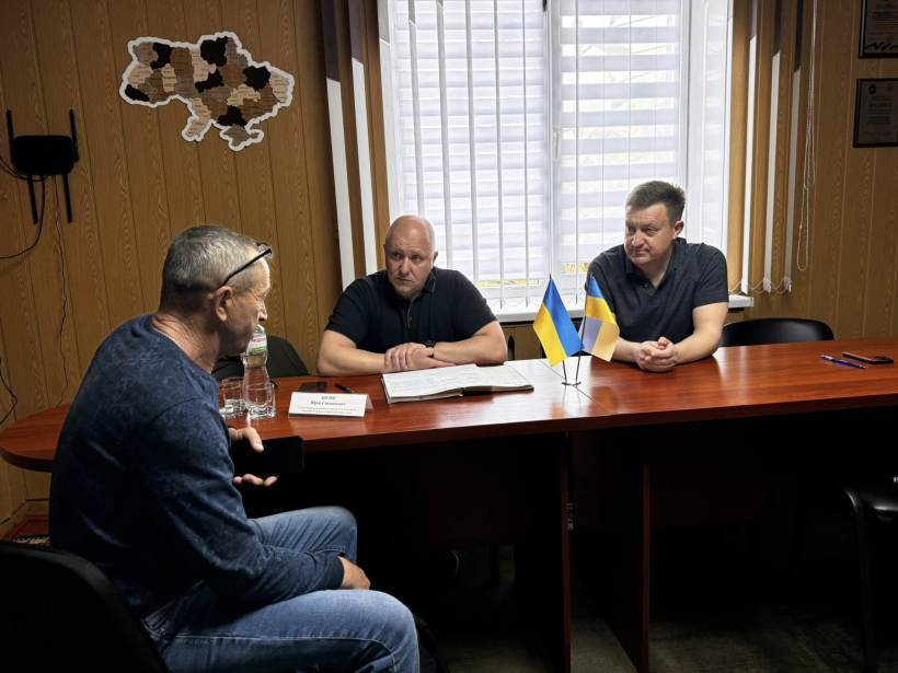 На фото зображені 2 чоловіки,що сидять за круглим столом, та житель Городоцької громади, який ділиться своїми проблемними питаннями. На фоні вікно, а на стіні висить карта України
