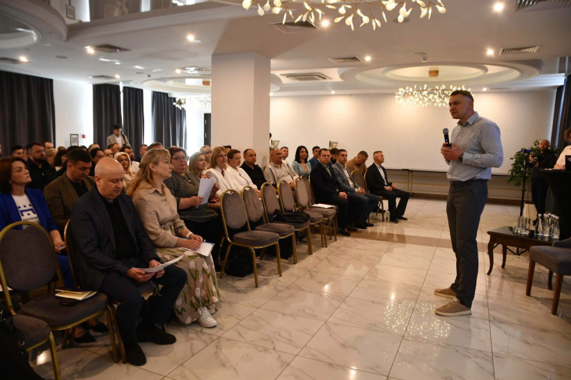 На фото зображено групу людей, що складається з чоловіків та жінок, а також спікера, який виступає,як доповідач. Зала зображена у пастельних тонах.