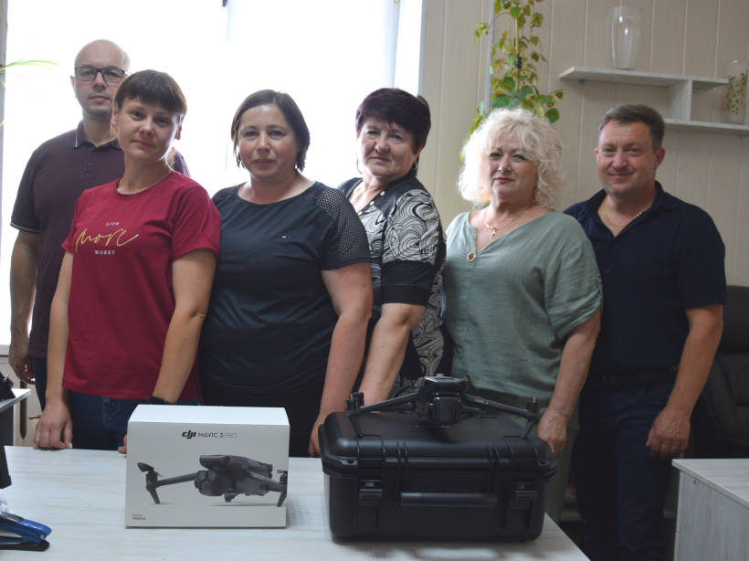 На знімку група людей стоїть у приміщенні біля столу, на якому розміщено два квадрокоптери в упаковках: DJI Mavic 3 PRO у білому коробі та ще один дрон у чорному кейсі. Це, ймовірно, представники Городоцької територіальної громади, які долучилися до передачі сучасного обладнання для потреб Збройних Сил України. Усі учасники фото виглядають зосередженими та небайдужими.