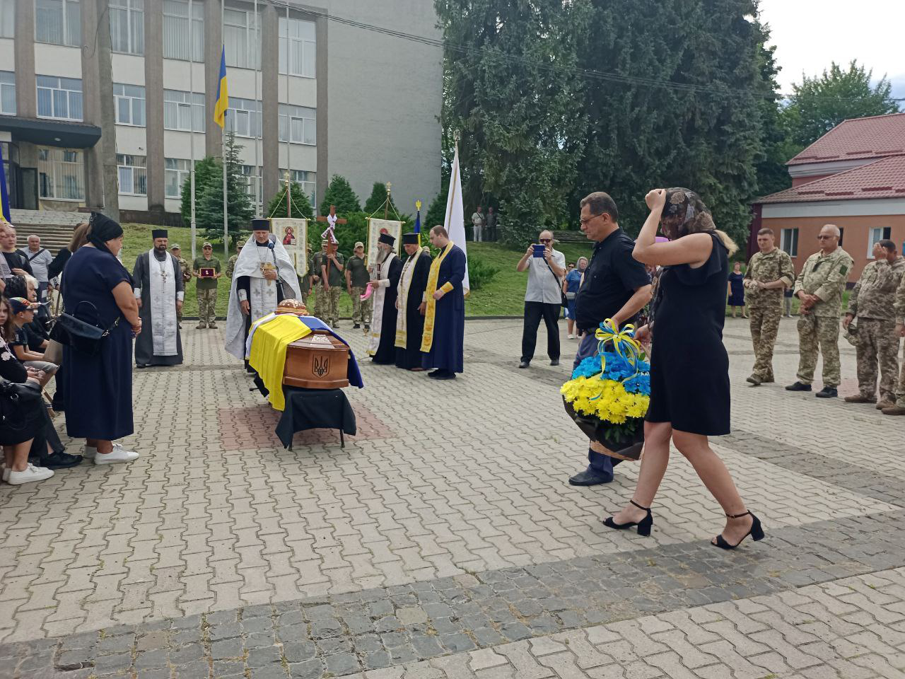 На головній площі міста Острог міський голова покладає кошик квітів до труни полеглого Героя Дениса Ціося. На задньому плані біля труни стоять п'ятеро священників, військові, рідні воїна та містяни.