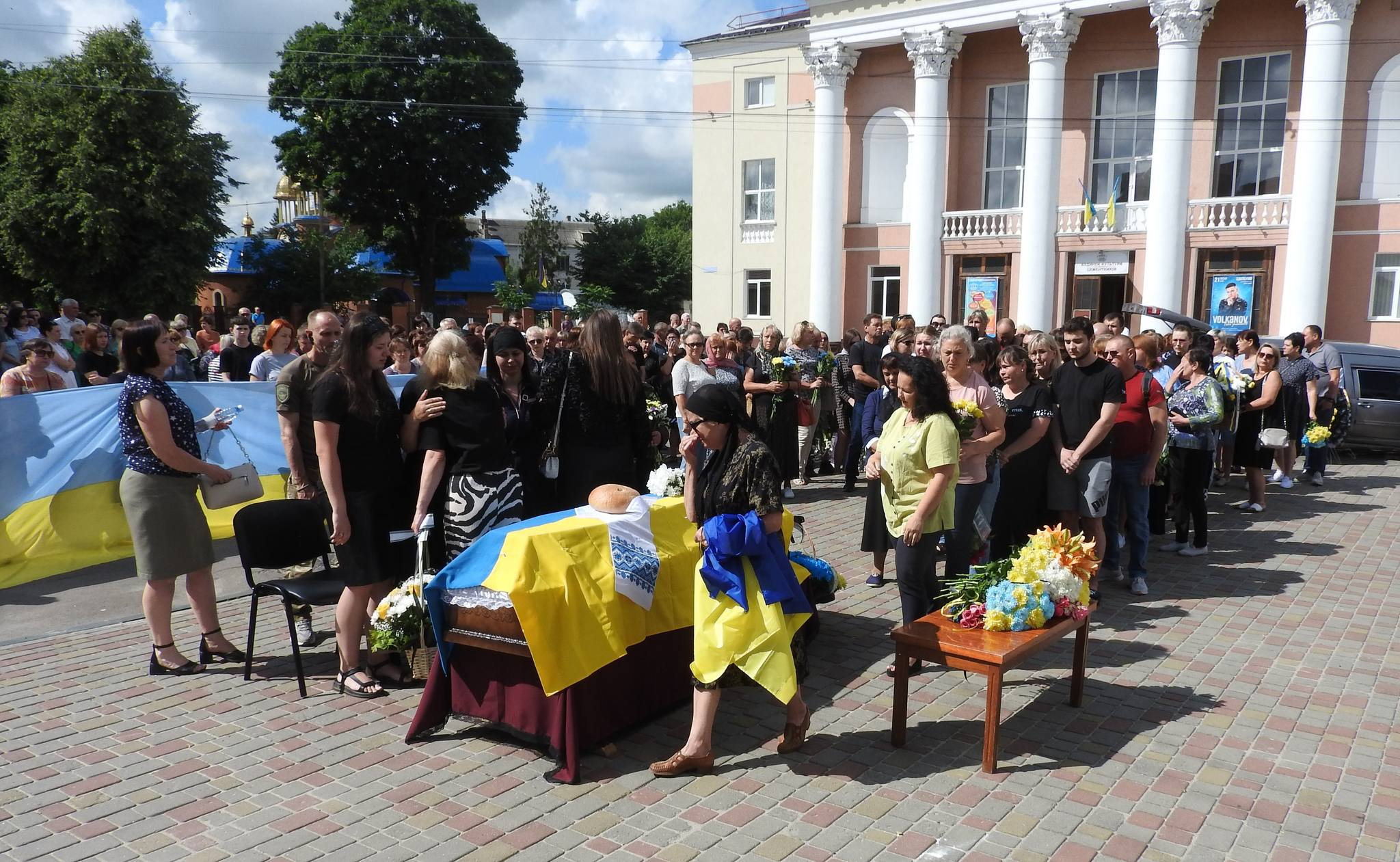 На площі перед будинком культури стоїть труна накрита жовто-блакитним прапором до якої підходять люди щоб попрощатися та покласти квіти. На площі багато людей, дехто плаче.