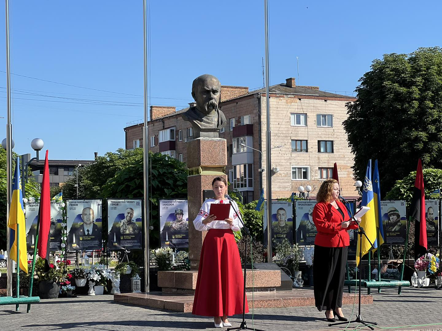 На площі перед пам'ятником Тарасу Шевченку стоять святково одягнені дві ведучі свята. За ними розміщені банери з портретами полеглих у війні воїнів, біля яких багато квітів, поруч жовто-блакитний прапор.