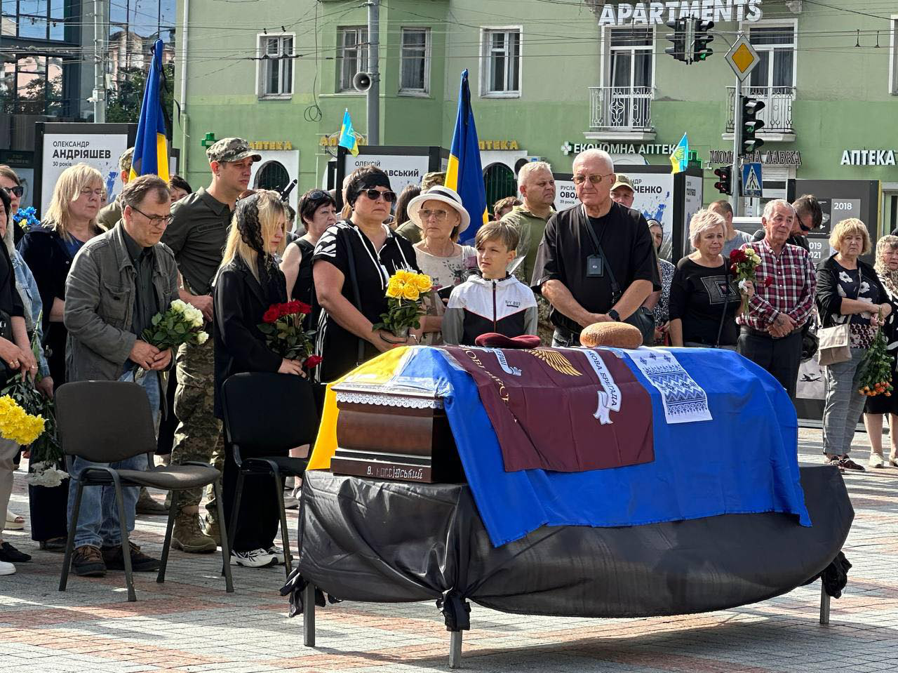 На майдані Незалежності міста Рівне стоїть труна накрита жовто-блакитним прапором. Поруч стоять люди в чорному одязі та квітами в руках, вони засмучені.