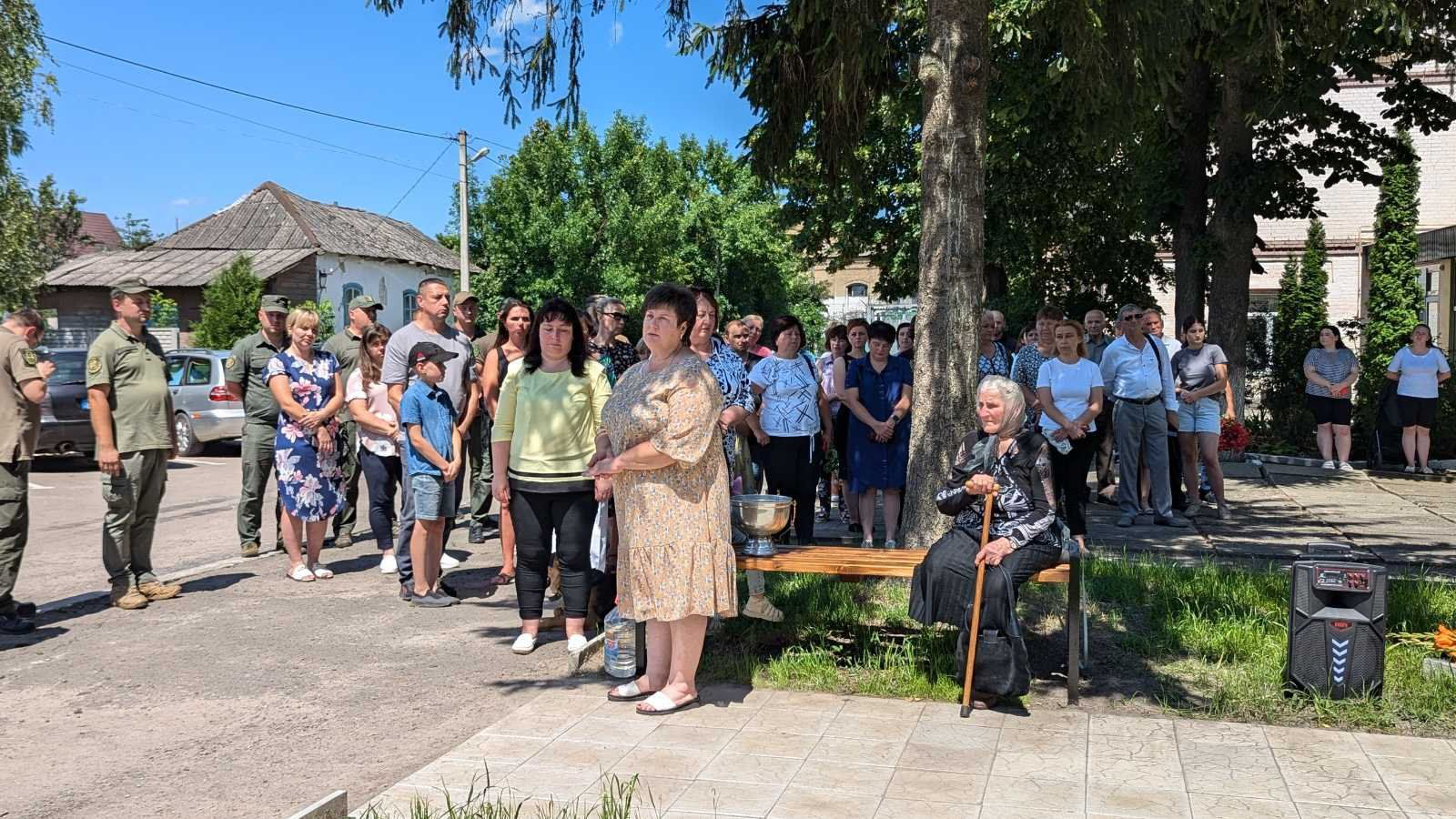 Білокриницький сільський голова Тетяна Гончарук звертається до мешканців села Городище з промовою. Місцеві її слухають, старенька бабуся з паличкою сидить на лавці поруч Алеї, засмучено слухає очільника громади з похиленою головою.