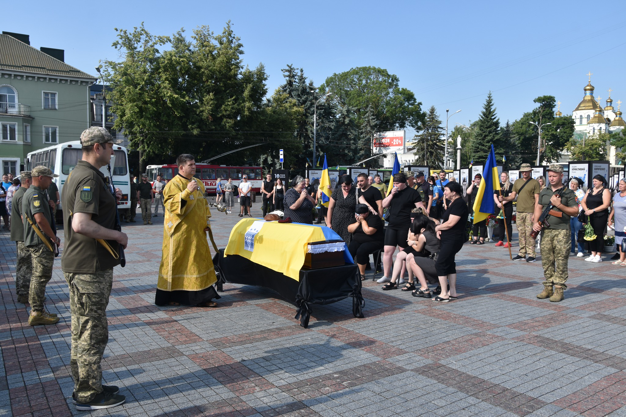 Православний батюшка відправляє молебнь біля труни полеглого воїна. Поруч рідні у чорному, які плачуть. За ними люди, які прийшли провести воїна в останню дорогу.