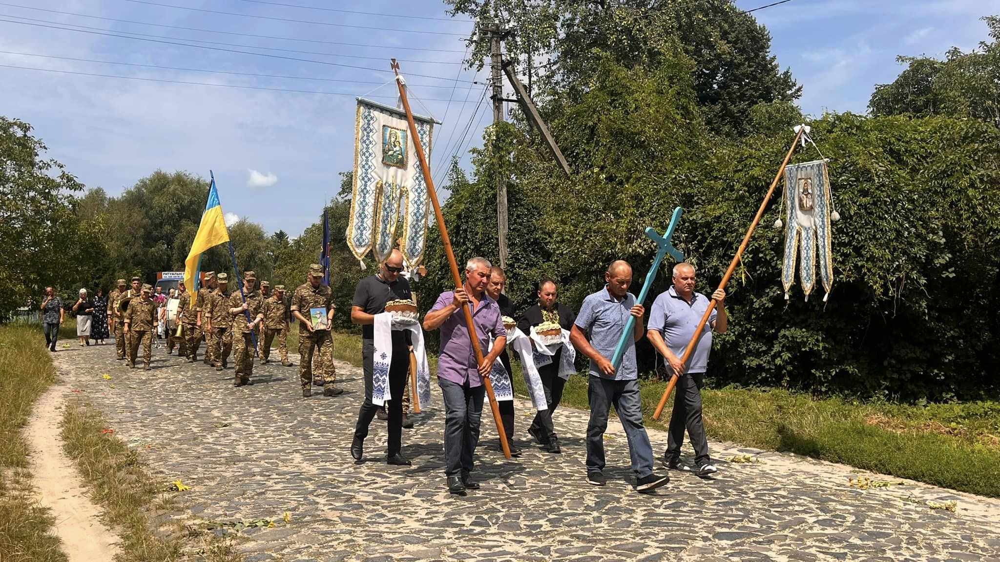 Йде поховальна процесія, чоловіки несуть у руках хрести, два короваї на рушниках, що означає поховання неодруженого хлопця. Позаду йдуть військові, які несуть портрет полеглого Героя.