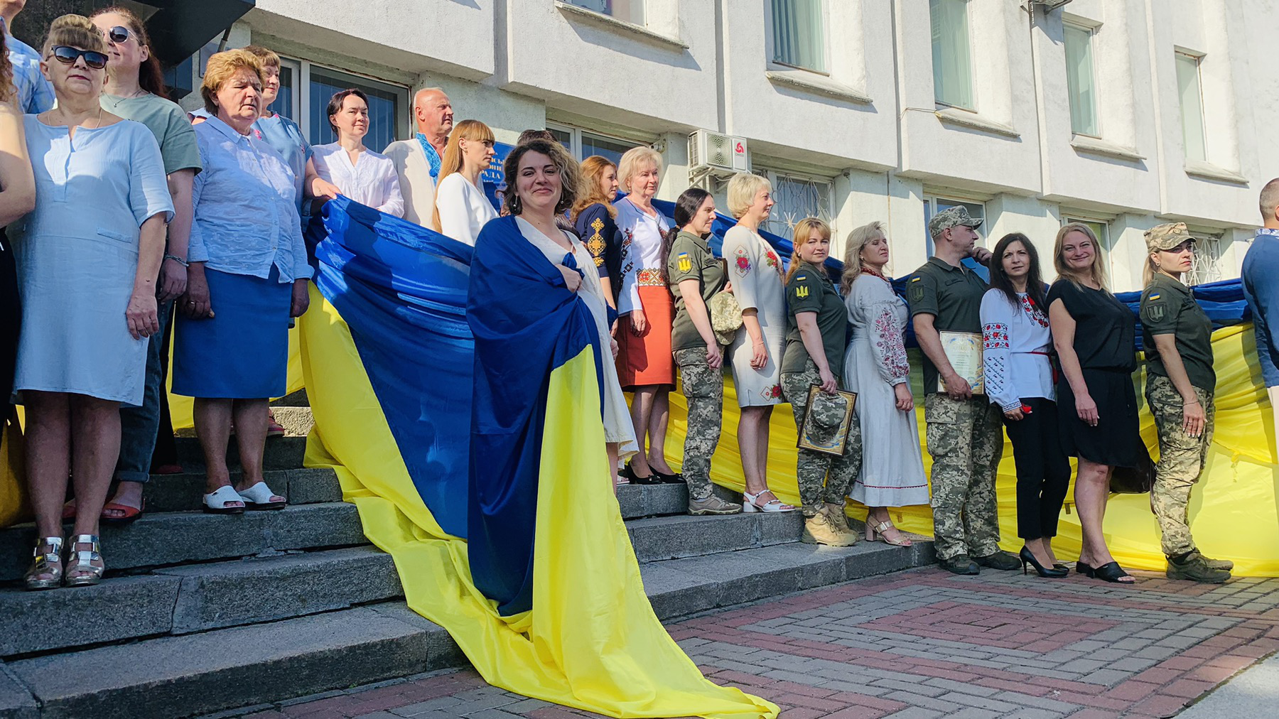 На вулиці перед адмінбудівлею Рівненської райдержадміністрації стоять святково одягнені працівники даної установи та військові і тримають жовто-блакитний прапор