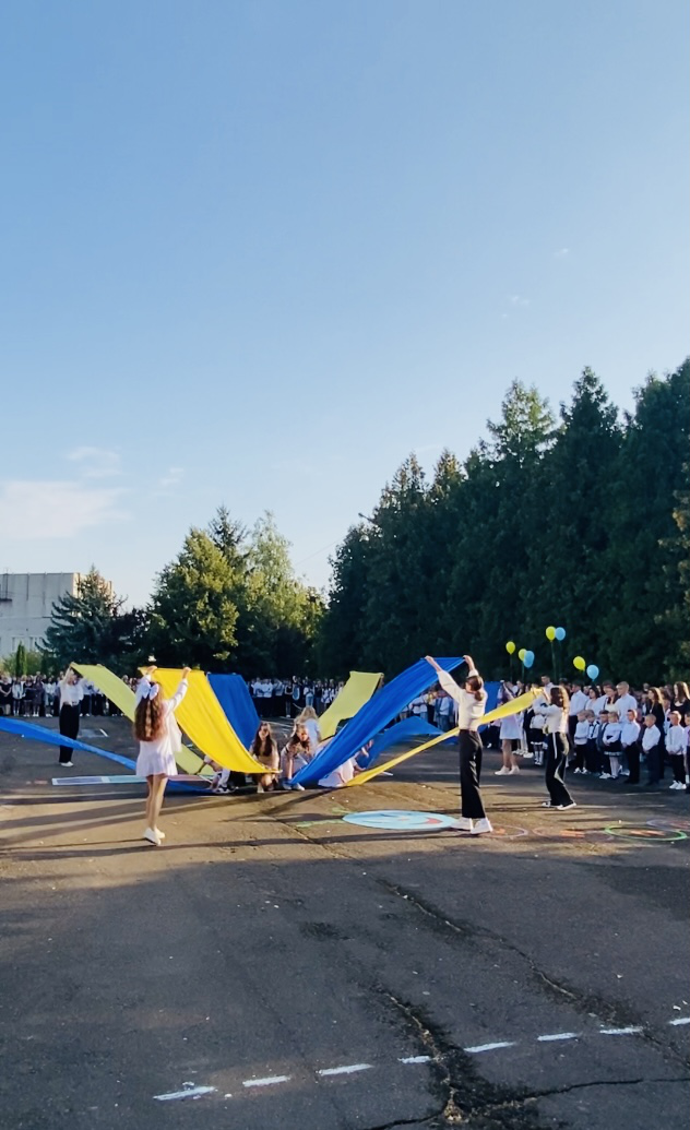 Учні на шкільному подвір'ї виконують танець з прапорами. Інші учні стоять на лінійці й спостерігають